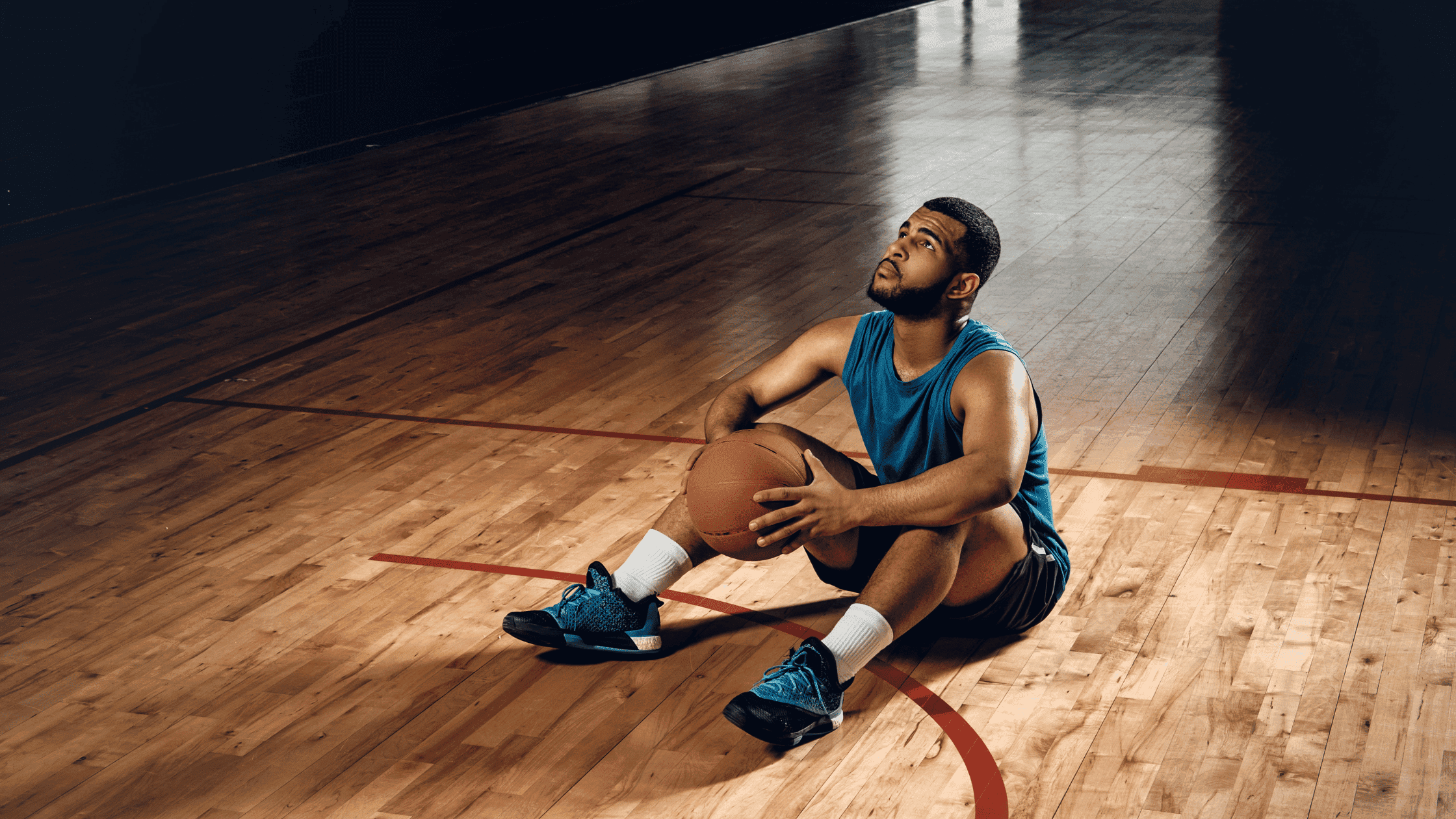 Second Team Basketball at Prep School The Pros and Cons (29) Young male basketball player sitting on the court holding a basketball, preparing for a shot, in a gym with polished wooden flooring.
