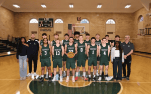 Deerfield Academy varsity basketball team poses at center court before tip-off, surrounded by coaches and staff under the scoreboard.