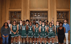 The Deerfield Academy boys basketball team poses together in uniform inside a trophy-lined hallway, celebrating their team unity.