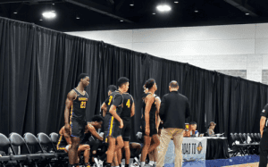 Massanutten players in black and gold jerseys gather near the bench during a timeout at a prep basketball showcase, with coaches and scorers’ table in view.