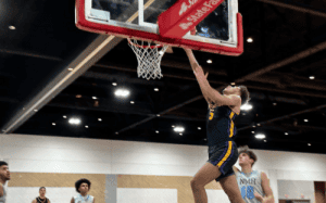 Massanutten guard in black drives and extends for a right-hand finish at the rim against NMH, with crowd, teammates, and ceiling lights in the background.