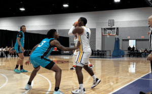 A Massanutten player in a white jersey receives the ball on the wing while a teal-uniform defender closes out at a prep showcase, with baseline and bench visible.