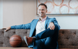 Alan Stein Jr. sitting on a couch in a suit with a basketball beside him, representing leadership and athlete performance coaching.