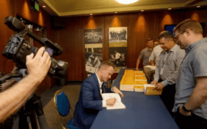 Alan Stein Jr. signing books at an event with attendees and a camera recording the moment.