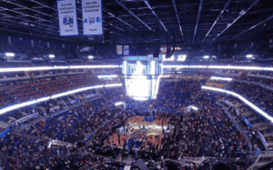 Wide view of an NBA basketball arena with a packed crowd and bright scoreboard.