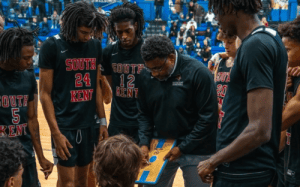 South Kent basketball coach Craig Carter giving instructions during a timeout huddle with players on the court.