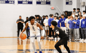 Blue Ridge School basketball player dribbles against a defender during a prep school game as teammates and fans watch from the sidelines.