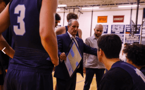Coach Brian Fruscio gives instructions to Canterbury School basketball players during a timeout, holding a clipboard and emphasizing strategy and teamwork.
