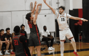 Hun School basketball player shoots over a defender during a home game against Blair Academy, with teammates and coaches watching courtside.