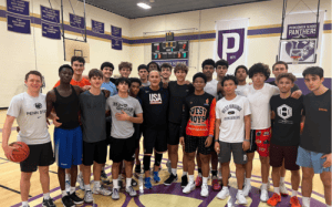 The Perkiomen School basketball team gathers around a USA Basketball coach during a high-energy gym training session, emphasizing teamwork and player development.