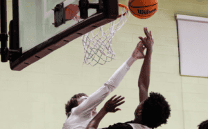 Prep school basketball defender contesting a shot at the rim as an offensive player finishes through contact in a high school gym.
