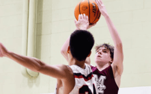 Prep school basketball player rising for a contested jump shot as a defender challenges at full extension inside a high school gym.